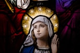 A stained glass depiction of a woman with a serene expression, surrounded by a halo of stars. She is wearing a white headscarf and blue robe against a richly colored background of deep reds and purples.