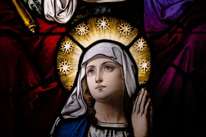A stained glass depiction of a woman with a serene expression, surrounded by a halo of stars. She is wearing a white headscarf and blue robe against a richly colored background of deep reds and purples.