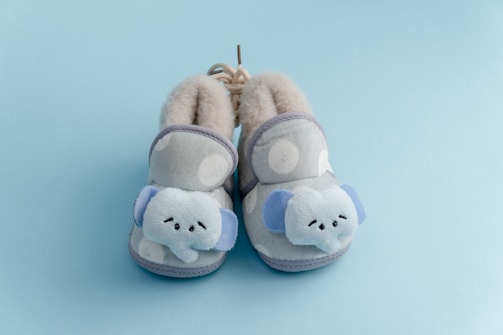 A pair of cozy, soft baby slippers featuring plush elephant faces on the front. The slippers are light gray with white polka dots and have a fluffy interior, set against a light blue background.