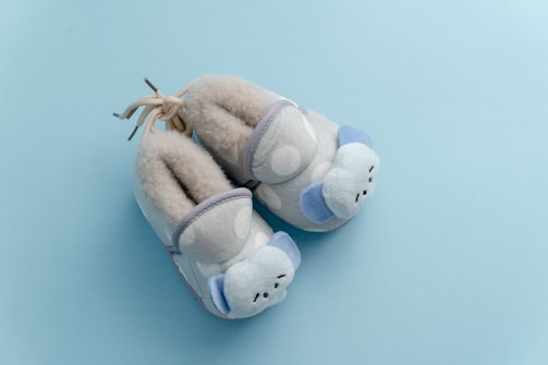 A pair of soft, plush baby slippers adorned with adorable animal faces, featuring shades of pastel blue and cream. The slippers are positioned on a light blue surface, suggesting a gentle and soothing atmosphere.