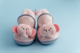 A pair of soft, pink, polka-dotted baby booties with elephant faces and ears on the front. The background is light blue, creating a gentle contrast with the booties. The booties have gray trim and soft, cushioned interiors.