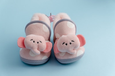 A pair of soft, pink, polka-dotted baby booties with elephant faces and ears on the front. The background is light blue, creating a gentle contrast with the booties. The booties have gray trim and soft, cushioned interiors.
