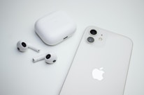 A pair of wireless earbuds with a charging case are placed next to a smartphone. Both the case and the phone are white, and the smartphone features dual cameras. The image has a minimalistic and clean aesthetic.