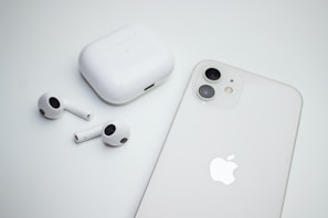 A pair of wireless earbuds with a charging case are placed next to a smartphone. Both the case and the phone are white, and the smartphone features dual cameras. The image has a minimalistic and clean aesthetic.