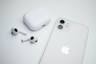 A pair of wireless earbuds with a charging case are placed next to a smartphone. Both the case and the phone are white, and the smartphone features dual cameras. The image has a minimalistic and clean aesthetic.