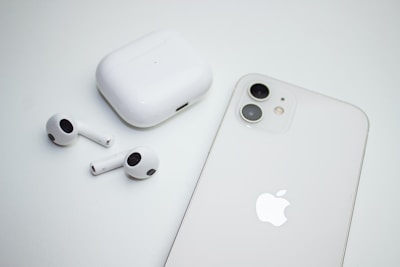 A pair of wireless earbuds with a charging case are placed next to a smartphone. Both the case and the phone are white, and the smartphone features dual cameras. The image has a minimalistic and clean aesthetic.