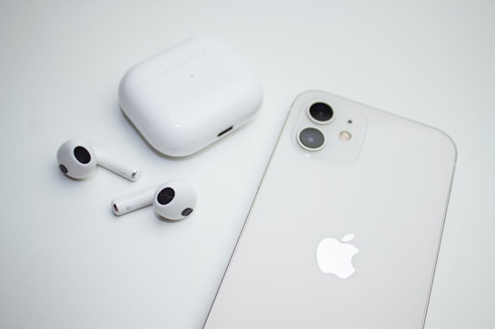 A pair of wireless earbuds with a charging case are placed next to a smartphone. Both the case and the phone are white, and the smartphone features dual cameras. The image has a minimalistic and clean aesthetic.
