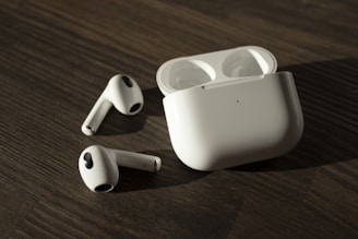A pair of wireless earphones with their charging case placed on a wooden surface. The earphones and case are white, with the earphones lying outside the open case.