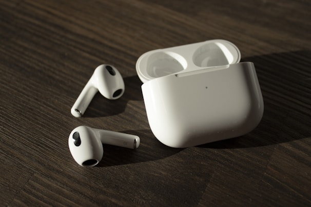 A pair of wireless earphones with their charging case placed on a wooden surface. The earphones and case are white, with the earphones lying outside the open case.
