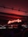 A dimly lit bar setting featuring a prominent neon sign reading 'Aperitivo Bar' in bright red. Shelves stocked with various bottles of liquor are visible beneath the sign. The lighting creates a warm and inviting atmosphere, with a person silhouetted in the foreground.
