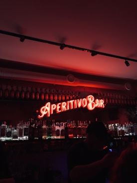 A dimly lit bar setting featuring a prominent neon sign reading 'Aperitivo Bar' in bright red. Shelves stocked with various bottles of liquor are visible beneath the sign. The lighting creates a warm and inviting atmosphere, with a person silhouetted in the foreground.
