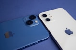 Two smartphones are placed side by side on a flat surface, one is blue and the other is white. Each device has dual cameras visible on the back, along with the manufacturer's logo.