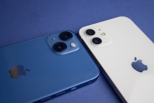 Two smartphones are placed side by side on a flat surface, one is blue and the other is white. Each device has dual cameras visible on the back, along with the manufacturer's logo.