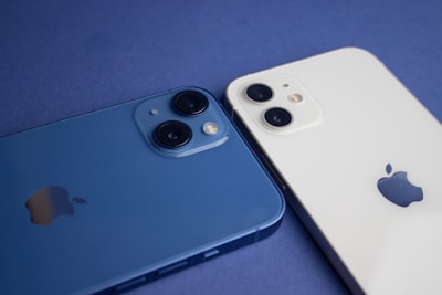 Two smartphones are placed side by side on a flat surface, one is blue and the other is white. Each device has dual cameras visible on the back, along with the manufacturer's logo.