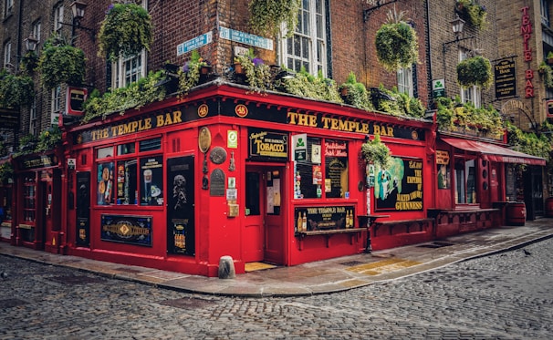A traditional Irish pub with a bright red exterior, adorned with various signs and advertisements. Hanging plants are draped across the facade, adding greenery to the scene. The cobbled street adds to the quaint and historic ambiance, and the windows display an array of posters and decorative elements.