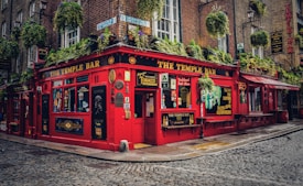 A traditional Irish pub with a bright red exterior, adorned with various signs and advertisements. Hanging plants are draped across the facade, adding greenery to the scene. The cobbled street adds to the quaint and historic ambiance, and the windows display an array of posters and decorative elements.