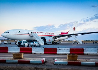 An Air Algerie airplane is parked on the tarmac at an airport. The aircraft is white with red accents and text. There are mobile staircases positioned at the aircraft’s doors and several vehicles parked nearby, including a few cars and a van. The scene includes rectangular red and white barriers lining part of the perimeter.