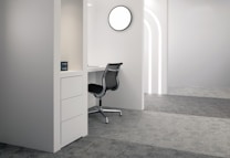 A modern, minimalist office space with a white desk, black office chair, and a set of drawers. The environment is well-lit with a circular light fixture on the wall. The surrounding area features grey carpeting and curved light strips on the walls, giving a clean and contemporary look.