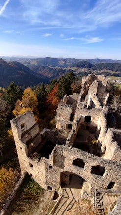 An aerial view of an ancient, crumbling stone ruin surrounded by dense forest with autumn-colored foliage. The structure's walls have multiple openings and arches, overlooking a vast landscape of rolling hills and distant mountains under a partly cloudy sky.