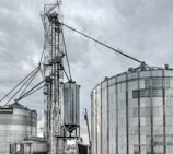 A large industrial scene featuring multiple metal silos and storage tanks set against a cloudy sky. The structures are interconnected by various walkways and pipes. The image highlights a complex network of steel frameworks and cylindrical storage units.
