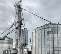 A large industrial scene featuring multiple metal silos and storage tanks set against a cloudy sky. The structures are interconnected by various walkways and pipes. The image highlights a complex network of steel frameworks and cylindrical storage units.