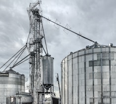 A large industrial scene featuring multiple metal silos and storage tanks set against a cloudy sky. The structures are interconnected by various walkways and pipes. The image highlights a complex network of steel frameworks and cylindrical storage units.
