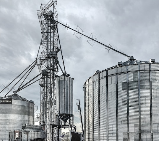 A large industrial scene featuring multiple metal silos and storage tanks set against a cloudy sky. The structures are interconnected by various walkways and pipes. The image highlights a complex network of steel frameworks and cylindrical storage units.