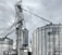 A large industrial scene featuring multiple metal silos and storage tanks set against a cloudy sky. The structures are interconnected by various walkways and pipes. The image highlights a complex network of steel frameworks and cylindrical storage units.