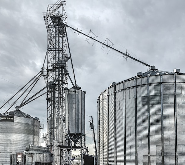 A large industrial scene featuring multiple metal silos and storage tanks set against a cloudy sky. The structures are interconnected by various walkways and pipes. The image highlights a complex network of steel frameworks and cylindrical storage units.