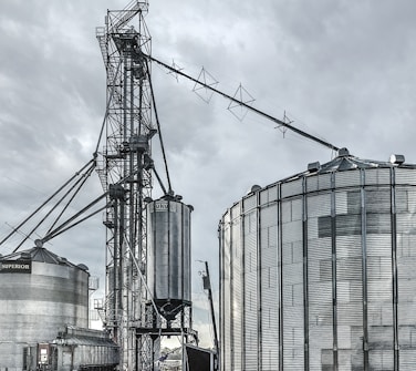 A large industrial scene featuring multiple metal silos and storage tanks set against a cloudy sky. The structures are interconnected by various walkways and pipes. The image highlights a complex network of steel frameworks and cylindrical storage units.