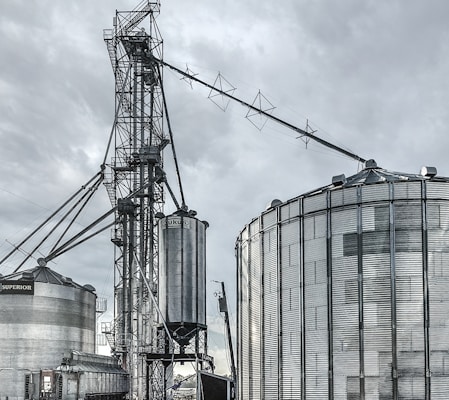 A large industrial scene featuring multiple metal silos and storage tanks set against a cloudy sky. The structures are interconnected by various walkways and pipes. The image highlights a complex network of steel frameworks and cylindrical storage units.