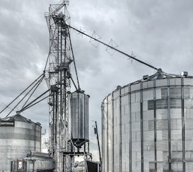 A large industrial scene featuring multiple metal silos and storage tanks set against a cloudy sky. The structures are interconnected by various walkways and pipes. The image highlights a complex network of steel frameworks and cylindrical storage units.