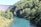 A river flows through a lush, forested area with dense green trees on its banks. To the left, a large concrete dam wall is visible, contrasting with the natural surroundings. Sunlight dapples through the foliage, reflecting off the clear blue-green water.