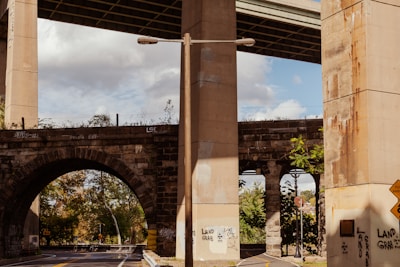 A scene under a bridge or overpass with large concrete pillars and an archway. The structure displays various forms of graffiti, including the phrase 'LAND GRAB.' Trees and foliage can be seen in the background through the arch. The sky is partly cloudy, creating a neutral yet slightly dramatic ambiance.