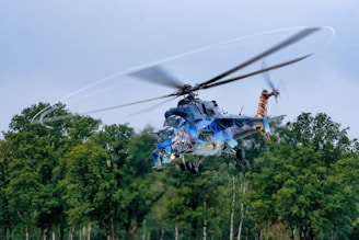 A helicopter is flying above a forested area with trees visible below. The helicopter is painted with vibrant colors and artistic designs, including what appears to be a tiger motif. The rotors are spinning rapidly, creating a blurred effect against the clear sky.