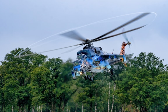 A helicopter is flying above a forested area with trees visible below. The helicopter is painted with vibrant colors and artistic designs, including what appears to be a tiger motif. The rotors are spinning rapidly, creating a blurred effect against the clear sky.