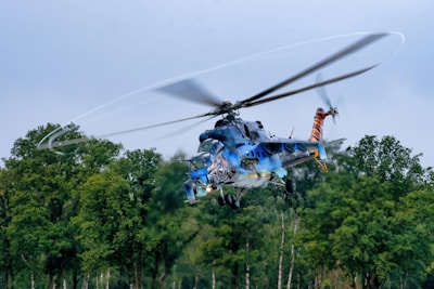 A helicopter is flying above a forested area with trees visible below. The helicopter is painted with vibrant colors and artistic designs, including what appears to be a tiger motif. The rotors are spinning rapidly, creating a blurred effect against the clear sky.
