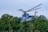 A helicopter is flying above a forested area with trees visible below. The helicopter is painted with vibrant colors and artistic designs, including what appears to be a tiger motif. The rotors are spinning rapidly, creating a blurred effect against the clear sky.