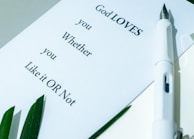 A white paper with text saying 'God LOVES you Whether you Like it OR Not' rests on a surface. A white pen lies next to the paper, and there are green leaves in the corner.