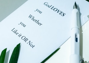 A white paper with text saying 'God LOVES you Whether you Like it OR Not' rests on a surface. A white pen lies next to the paper, and there are green leaves in the corner.