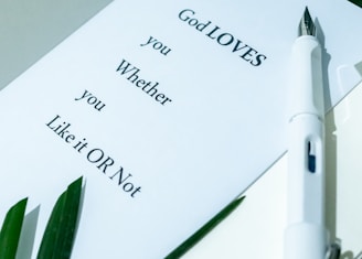 A white paper with text saying 'God LOVES you Whether you Like it OR Not' rests on a surface. A white pen lies next to the paper, and there are green leaves in the corner.