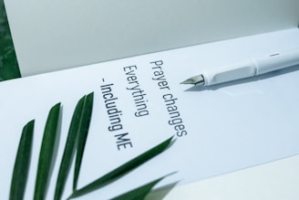 A white fountain pen lies on an open notebook page containing the inspirational text 'Prayer changes everything - including me'. A few green leaves are placed beside the text, adding a touch of nature to the composition.