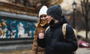 A young woman and man dressed in winter clothing walk together outdoors. Both are wearing warm jackets and knitted hats. The woman has a white beanie and a brown coat, and the man is wearing a dark blue patterned hat with a black coat and a backpack. They are near a colorful mural, and in the background, trees and a lamp post are visible.