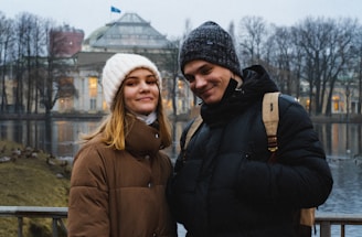 A young couple stands close together outdoors on a cold day. They are both warmly dressed in coats and knitted hats, smiling at the camera. Behind them is a large, ornate building with lights glowing through the windows, surrounded by water and winter trees.