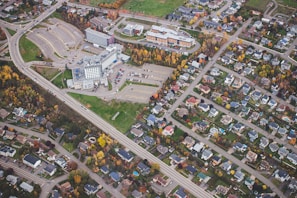 An aerial view capturing a suburban neighborhood. A large complex, possibly a hospital or office building, is surrounded by expansive parking lots. Residential houses with well-maintained yards line the streets. The trees show a mix of green and autumnal colors, adding vibrancy to the scene.
