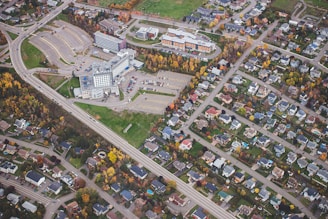 An aerial view capturing a suburban neighborhood. A large complex, possibly a hospital or office building, is surrounded by expansive parking lots. Residential houses with well-maintained yards line the streets. The trees show a mix of green and autumnal colors, adding vibrancy to the scene.