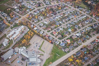 An aerial view of a suburban neighborhood showcasing a grid of residential houses with well-maintained yards. The scene includes a number of trees with autumn foliage scattered throughout the area. There is a significant presence of roads and a large parking lot near a set of modern buildings.