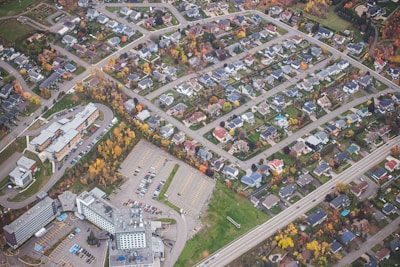 An aerial view of a suburban neighborhood showcasing a grid of residential houses with well-maintained yards. The scene includes a number of trees with autumn foliage scattered throughout the area. There is a significant presence of roads and a large parking lot near a set of modern buildings.