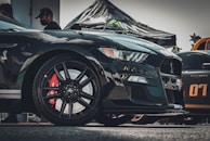 Close-up view of a sleek black sports car with a visible Shelby emblem and red Brembo brake calipers. The car is parked beside another car decorated with the number 07. In the background, there is a tent with visible text and some people wearing caps and masks.