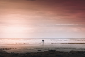 A solitary figure stands on a vast, empty beach facing the ocean, surrounded by a soft, warm light from the sky. The horizon line separates the calm, expansive sea from the dramatic, cloudy sky above, creating a serene and introspective scene.